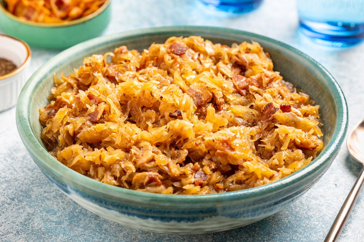 3/4th closeup shot of A ceramic bowl filled with cooked sauerkraut mixed with browned bacon pieces, set on a light blue surface with a spoon and small bowls in the background