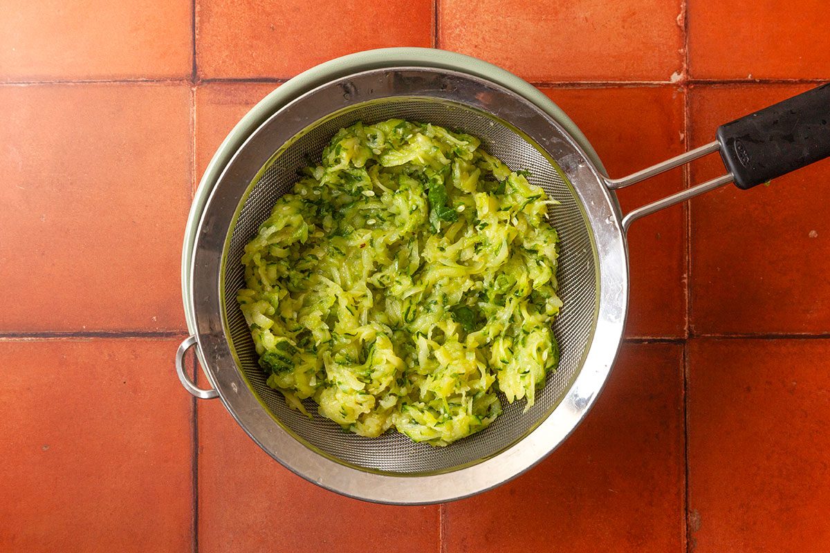 Overhead shot of grated zucchini mixed with green herbs in a metal strainer set over a bowl; set on a terracotta-tiled surface