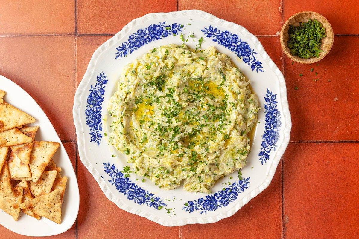 Overhead shot of smashed zucchini dip garnished with olive oil and fresh herbs, served on a patterned plate, with pita chips and a small bowl of chopped herbs on the side; set on a terracotta-tiled surface
