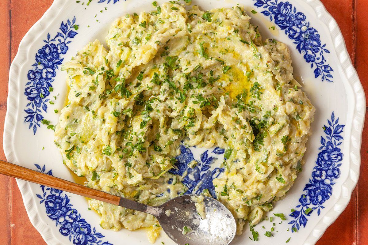 Close-up shot of smashed zucchini dip on a patterned plate; garnished with herbs and olive oil; set on a terracotta-tiled surface