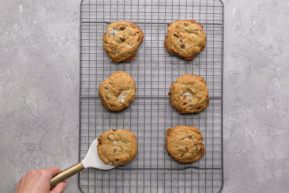 Six chocolate chip cookies are cooling on a metal wire rack over a gray surface. A hand using a spatula is about to lift one of the cookies from the rack.