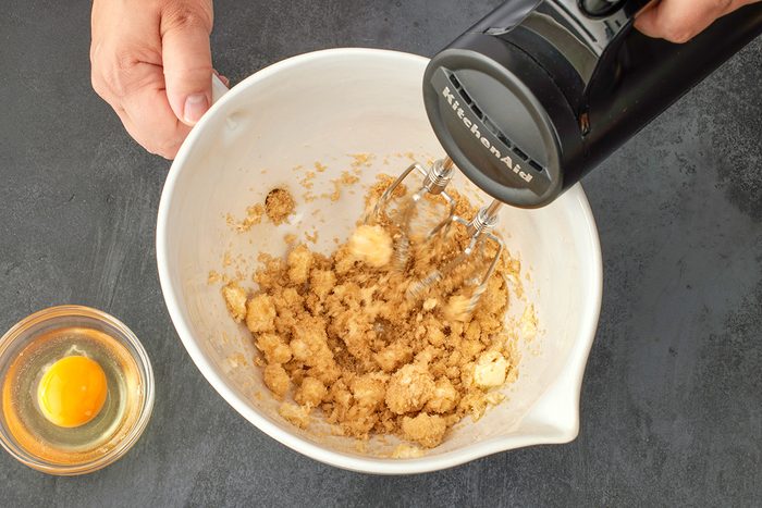 Overhead shot of a hand holding a white mixing bowl with sugar and butter being blended by a black electric mixer; while a small glass bowl containing a raw egg rests nearby on a dark countertop