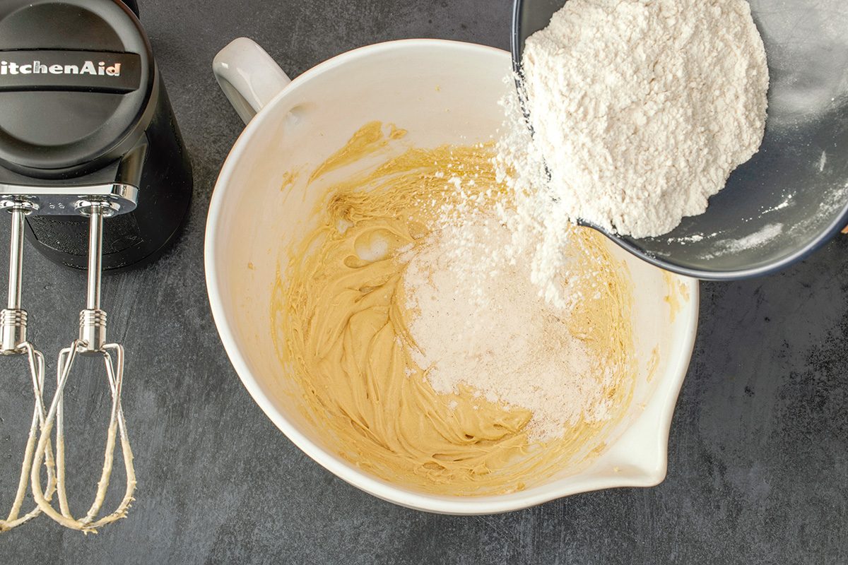 Overhead shot of a bowl of flour being poured into a mixing bowl with creamy batter; while an electric hand mixer with batter-coated beaters rests nearby on a dark countertop