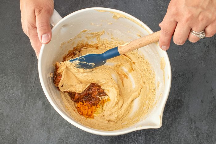 Overhead shot of hands holding a white mixing bowl filled with light brown batter; as a blue spatula stirs in dark brown and orange ingredients, all set on a black surface