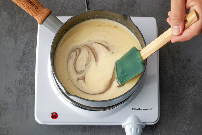 Overhead shot of a hand stirring a creamy mixture with a green spatula in a saucepan on a white electric stove; with visible swirls in the liquid