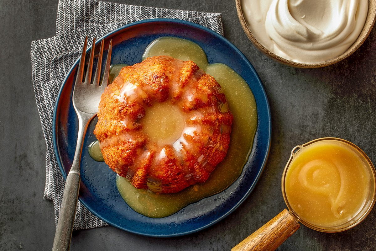 Overhead shot of a mini bundt cake topped with icing and sauce on a dark blue plate with a fork; placed beside a bowl of cream and a small pitcher of caramel sauce on a dark textured surface