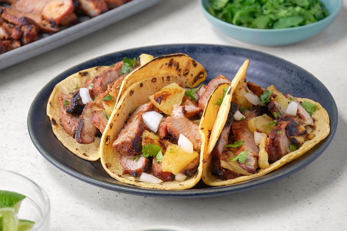 Three tacos filled with grilled meat, pineapple, diced onions, and cilantro are served on a dark plate, with a bowl of cilantro and a tray of extra meat visible in the background.