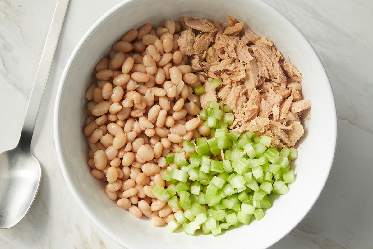 A white bowl contains three separated ingredients: white beans, flaked tuna, and chopped celery on a marble surface, with a spoon nearby.