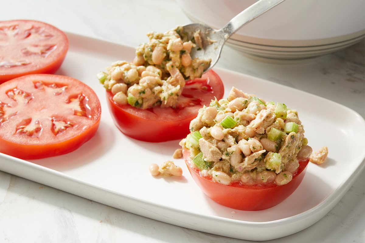 Two thick tomato slices on a white plate, one being topped with a spoonful of white bean salad mixed with diced celery, while the other remains plain.