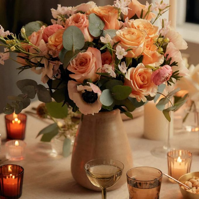 A ceramic vase filled with peach and pink roses, white flowers, and eucalyptus sits on a table surrounded by lit candles and a glass of white wine, creating a warm, romantic ambiance.