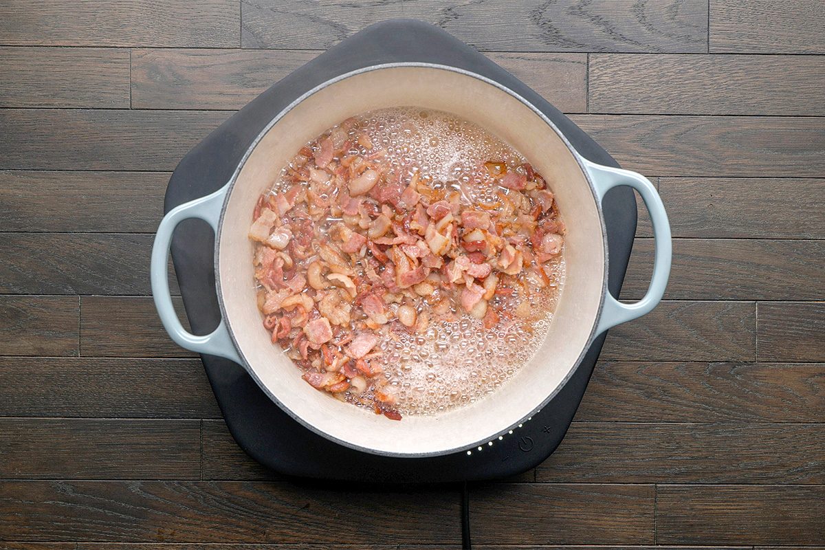 Chopped bacon pieces frying and sizzling in a round, light-colored pan on a black electric cooktop, placed on a dark wooden surface.