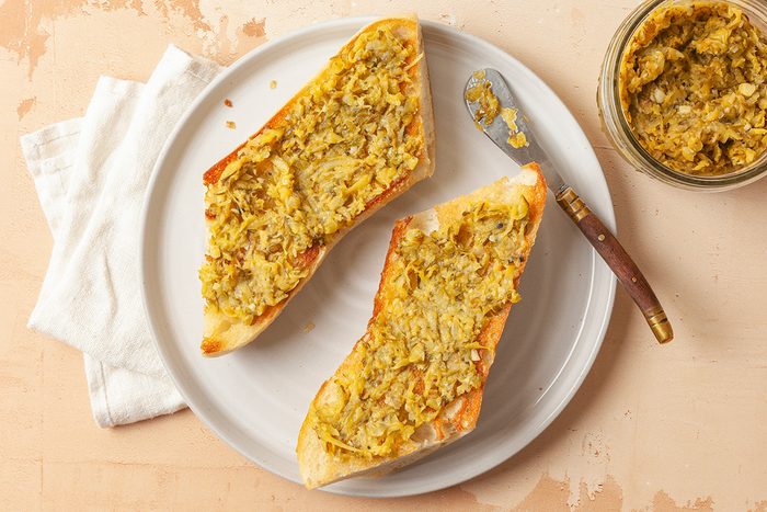 This is a close-up food shot; two toasted baguette slices with chunky yellow-green spread are on a white plate beside a matching jar and knife; all rest on light fabric over a beige surface