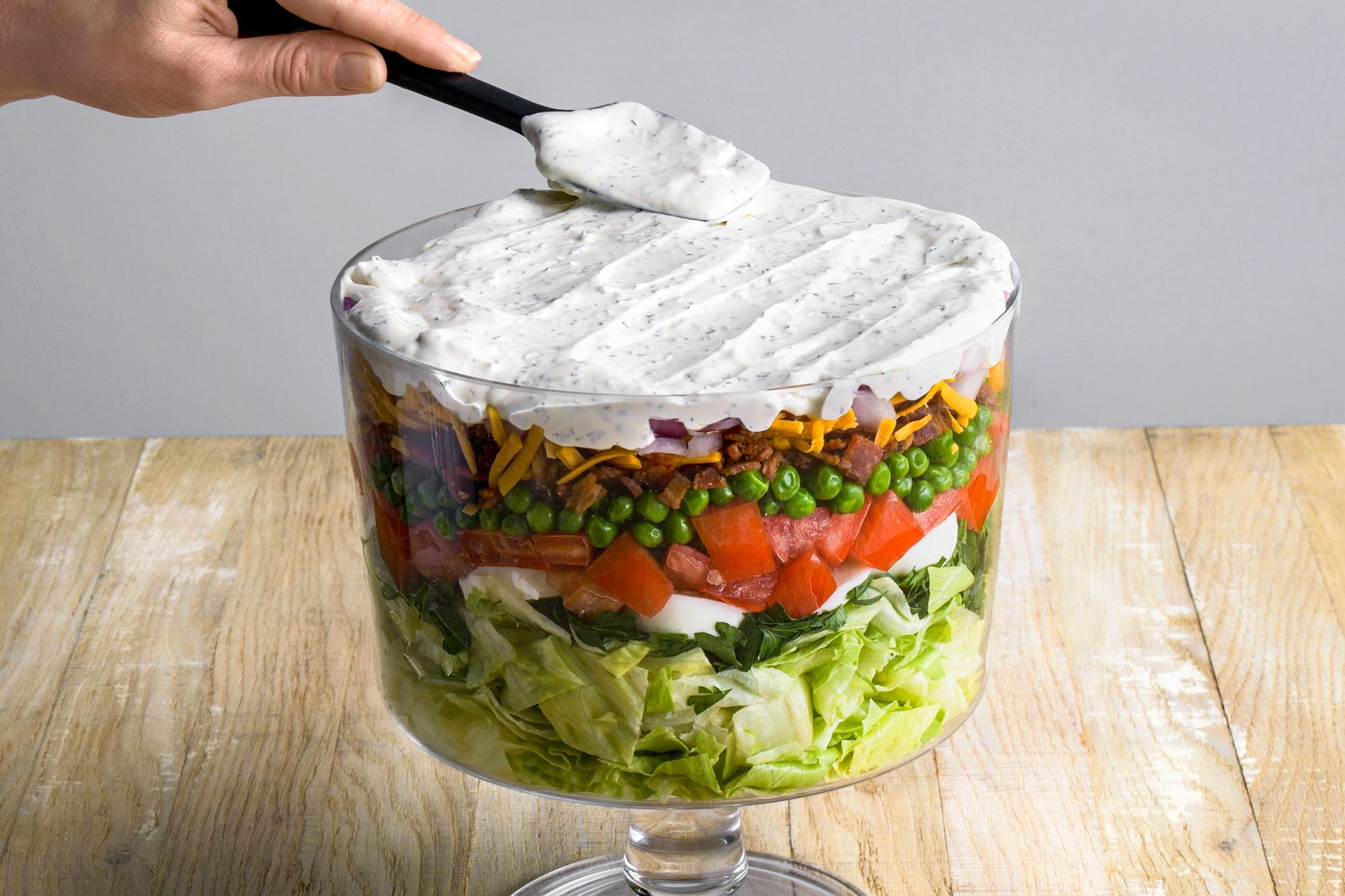 3/4 angle shot of a hand spreading creamy dressing over a layered salad in a clear glass bowl, showing layers of lettuce, spinach, tomatoes, peas, bacon, cheese, and onions;