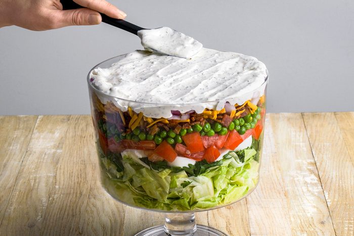 3/4 angle shot of a hand spreading creamy dressing over a layered salad in a clear glass bowl, showing layers of lettuce, spinach, tomatoes, peas, bacon, cheese, and onions;