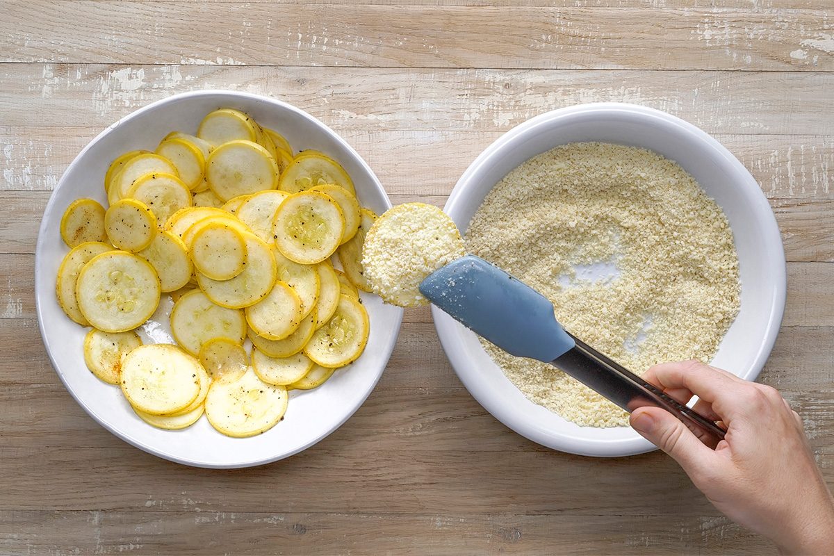 A hand uses tongs to dip a slice of yellow squash into a bowl of breading mixture, with a plate of sliced squash on the side, all on a wooden surface.
