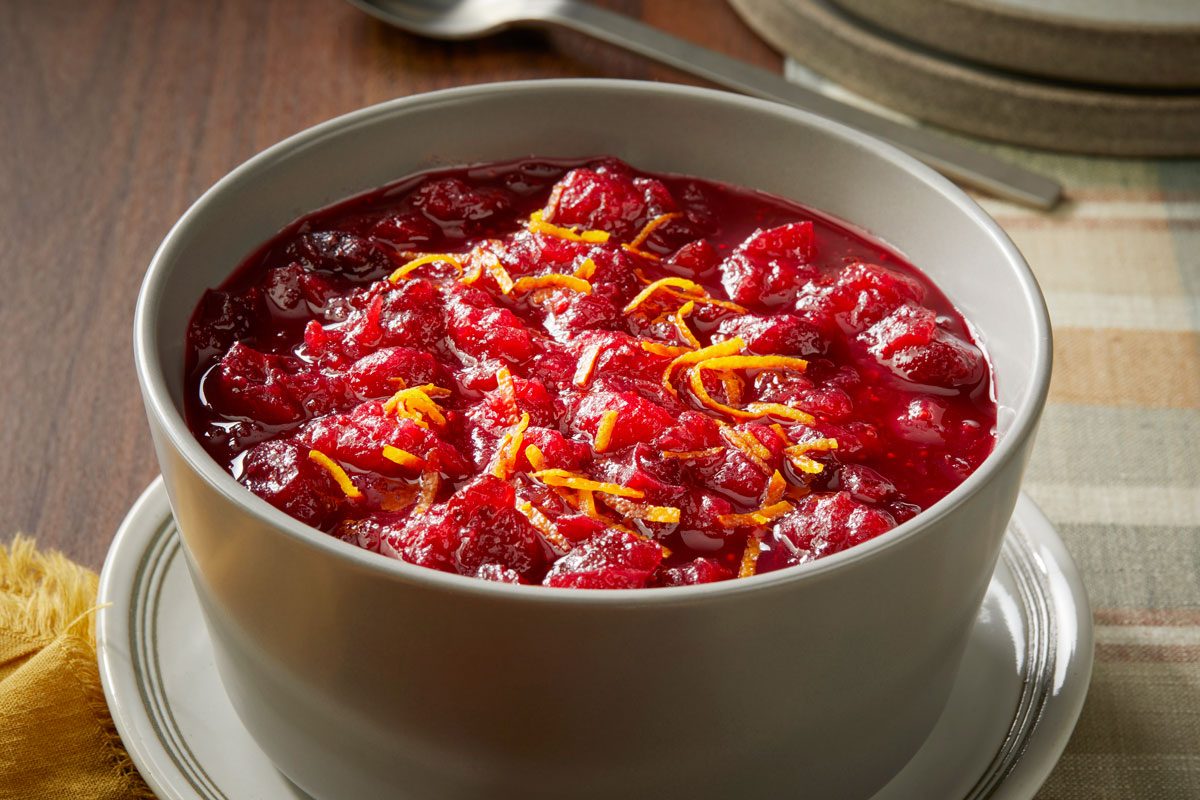 Close view shot of Aperol Spritz Cranberry Sauce served in a bowl, topped with orange zest, and set on a saucer over a checkered tablecloth, with plates and a spoon beside it;