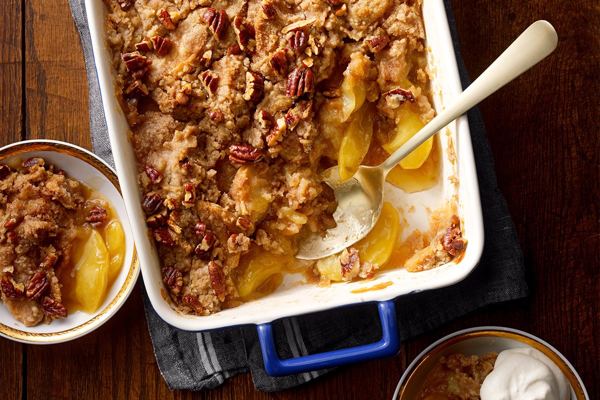 A baked apple crisp dessert in a white rectangular dish, topped with pecans and a crumbly streusel. A spoon rests inside, with servings in small bowls nearby on a wooden table.