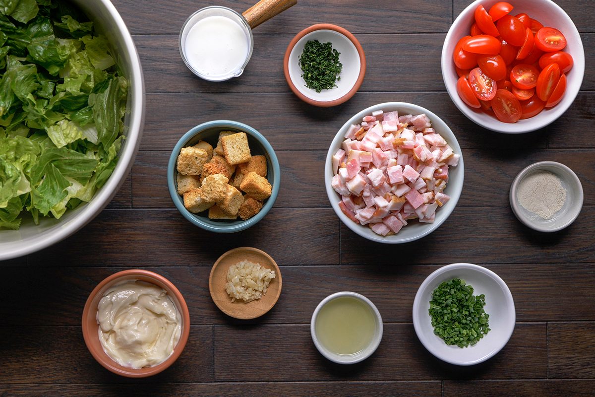Various salad ingredients, including chopped lettuce, cherry tomatoes, croutons, bacon bits, grated cheese, chives, Caesar dressing, lemon juice, and garlic, arranged in bowls on a dark wooden table.