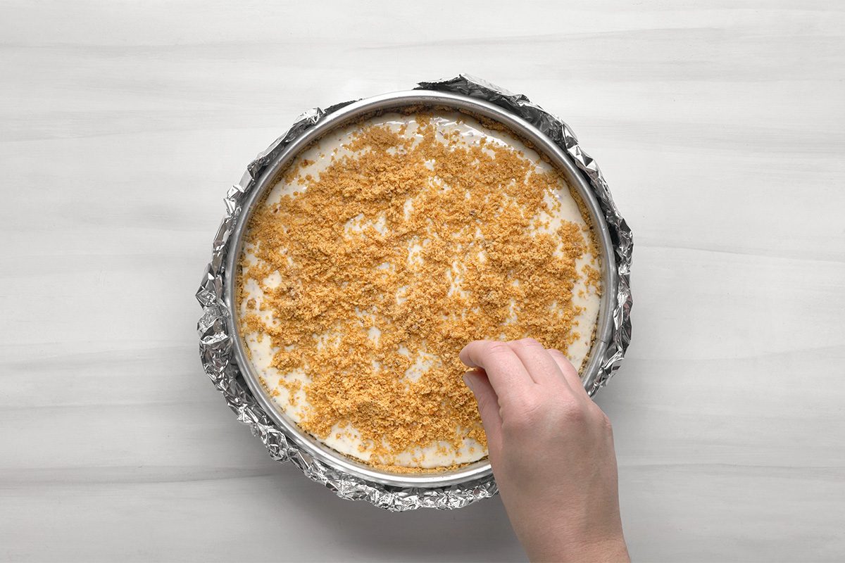 A hand sprinkles crumbs over a round cheesecake in a foil-wrapped pan, set on a light-colored surface.