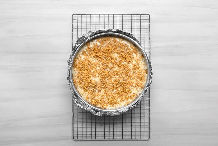 A round cheesecake in a foil-lined springform pan sits on a wire cooling rack. The cheesecake has a crumbly, golden topping and is viewed from above on a light-colored surface.