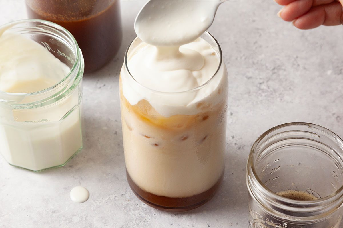 A hand is spooning creamy foam onto a glass of iced coffee. Surrounding the glass are jars containing milk, coffee, and syrup on a light-colored surface.