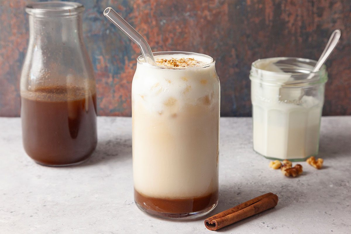 A glass of iced coffee with milk and a glass straw, topped with crushed nuts. Behind it are a jar of coffee syrup, a jar of cream or yogurt, and a cinnamon stick on the table.