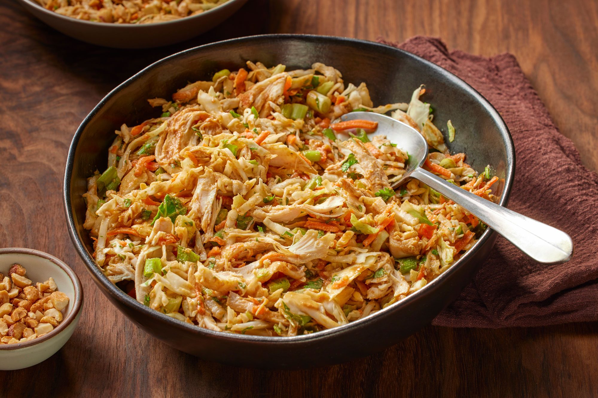 Close shot of Thai Chicken Coleslaw in a large bowl with cabbage, carrots, and green onions, with a silver spoon; chopped peanuts on the side, and a brown napkin nearby; all set on a wooden table;