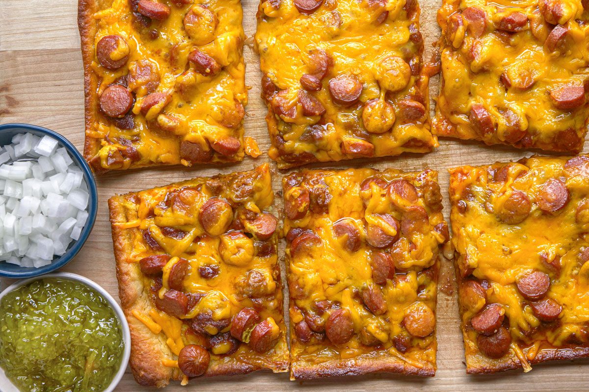 Overhead shot of Chili Dog Pizza; Rectangular slices of cheesy pizza topped with sliced sausages rest on a wooden board with small bowls of diced onions and green relish