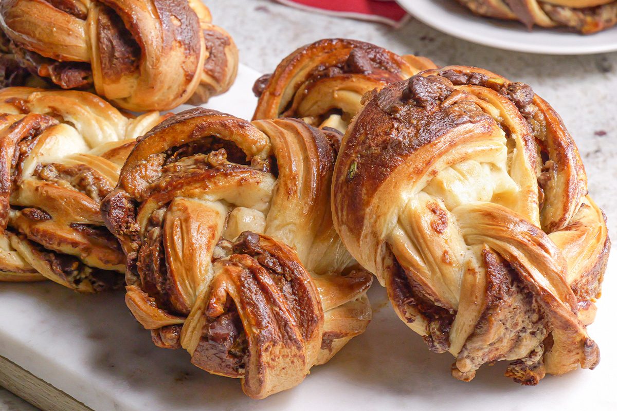 Close-up of three twisted pastries with a golden-brown, glossy crust and visible chocolate and cream cheese filling, displayed on a white marble surface.