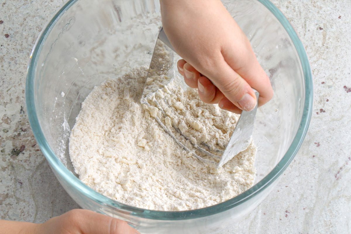 A person using a metal bench scraper to mix flour and butter in a clear glass bowl, preparing dough on a light-colored surface.