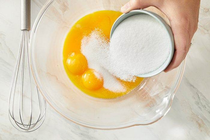A hand pours granulated sugar from a small bowl into a glass bowl containing three egg yolks on a marble countertop, with a whisk nearby.