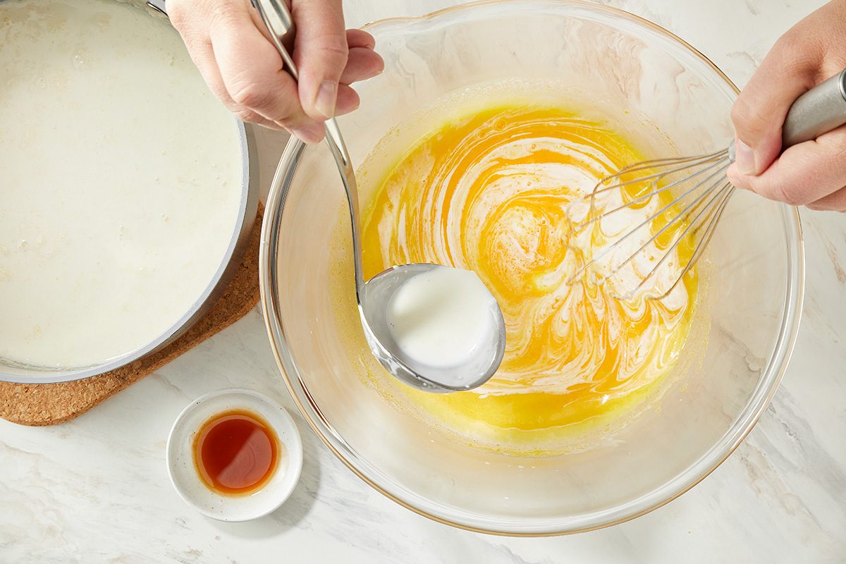 A person uses a ladle to pour milk into a bowl of beaten eggs while whisking. A small bowl of vanilla extract sits nearby on a white countertop.
