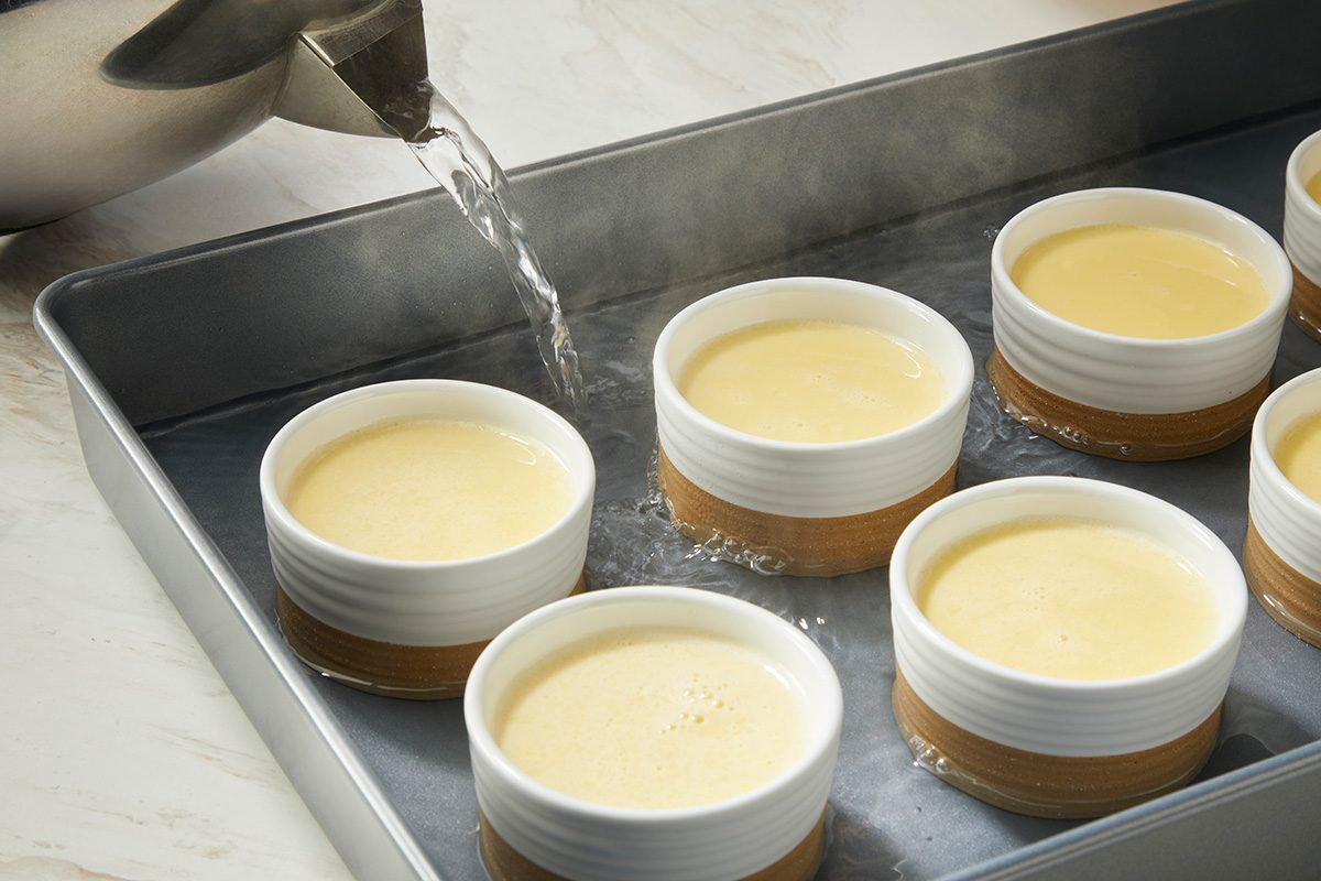 A metal kettle pours hot water into a baking pan holding several white ramekins filled with custard, preparing a water bath for baking.
