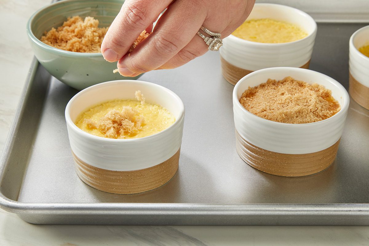 A hand sprinkles crumb topping onto individual ramekins filled with custard on a metal baking sheet, preparing a dessert.