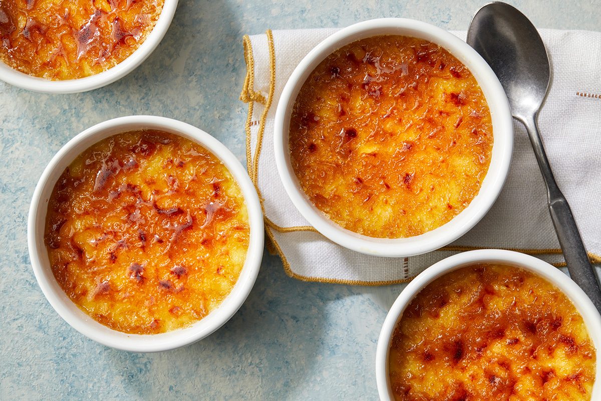 Four ramekins of crème brûlée with caramelized tops are arranged on a light blue surface, with a spoon placed on a folded napkin beside them.