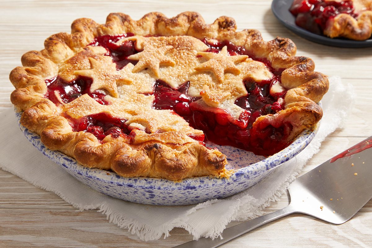 A cherry pie with a golden, star-shaped crust sits on a white cloth. One slice is missing, revealing the red cherry filling. A pie server and a plate with a slice of pie are in the background.