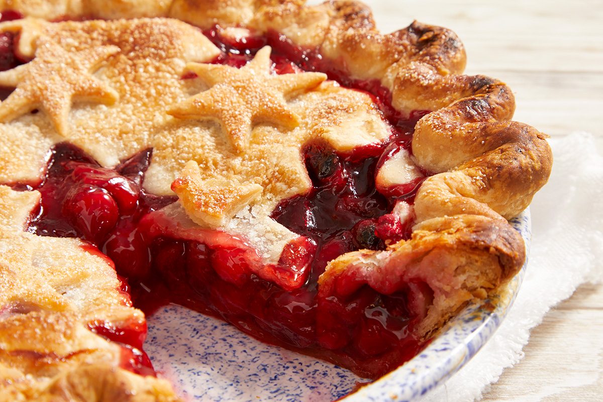 A close-up of a berry pie with a golden, flaky crust decorated with pastry stars. A slice has been removed, revealing the juicy, red berry filling inside. The pie sits on a white and blue dish.