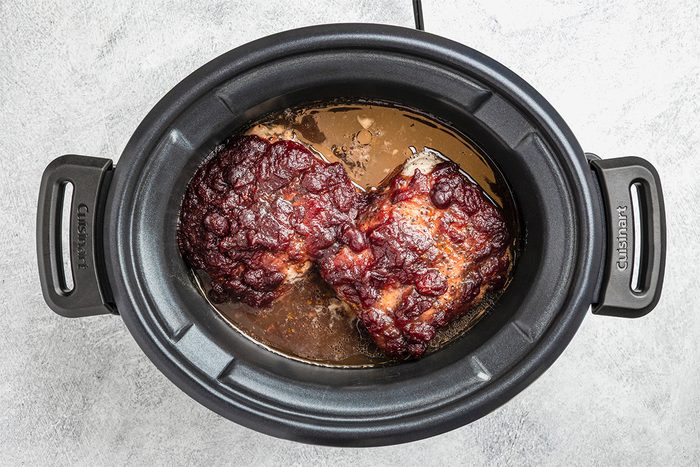 A slow cooker filled with two pieces of cooked meat covered in a dark, caramelized sauce, surrounded by juices, sitting on a light-colored countertop.