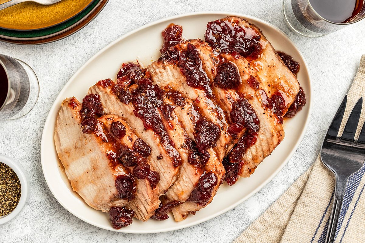 Sliced turkey breast topped with a chunky cranberry sauce arranged on an oval white plate, with a fork, knife, napkin, and glasses of red beverage nearby on a light surface.