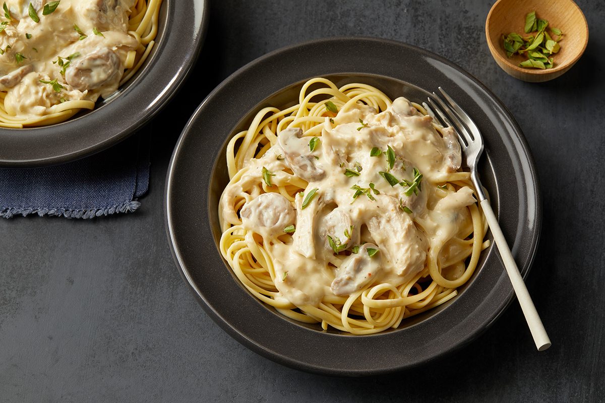 A plate of spaghetti topped with creamy white Alfredo sauce and garnished with chopped herbs, served on a dark plate with a fork. A small bowl of extra herbs is nearby.