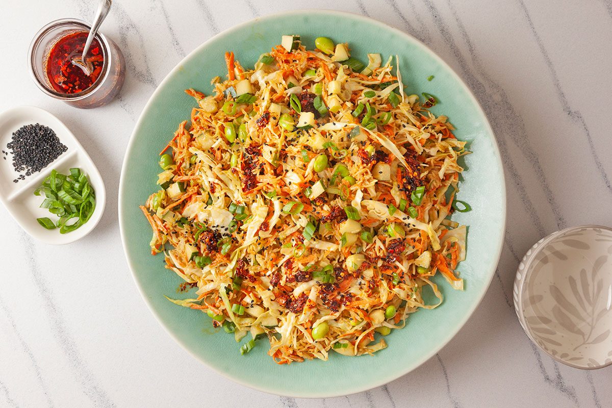 Overhead shot of Crispy Rice Salad on a large green plate, featuring shredded cabbage, carrots, and edamame, topped with chili flakes, sesame seeds, and chopped green onions; On the marble surface beside it are a small white dish with black sesame seeds and sliced green onions, along with a jar of chili oil and a spoon;