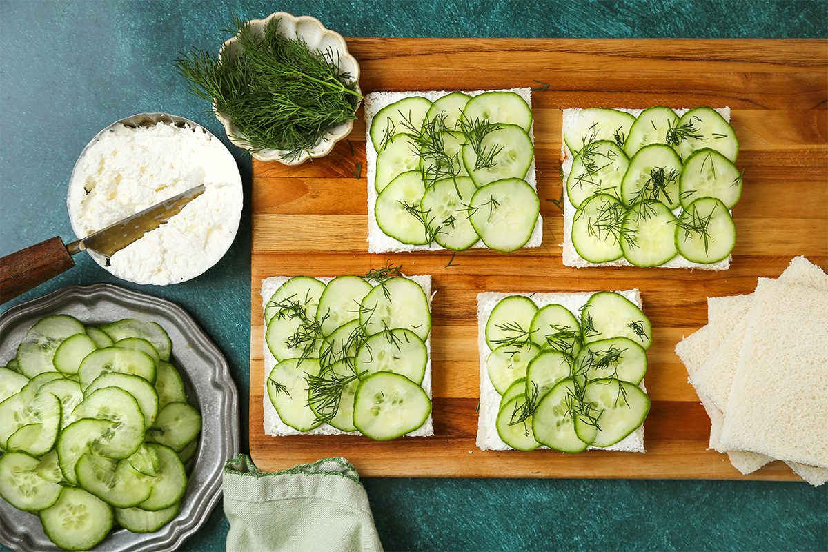 Four slices of bread topped with a creamy spread, cucumber slices, and fresh dill are arranged on a wooden board. A bowl of dill, a spreader, and extra cucumbers are nearby on a green background.