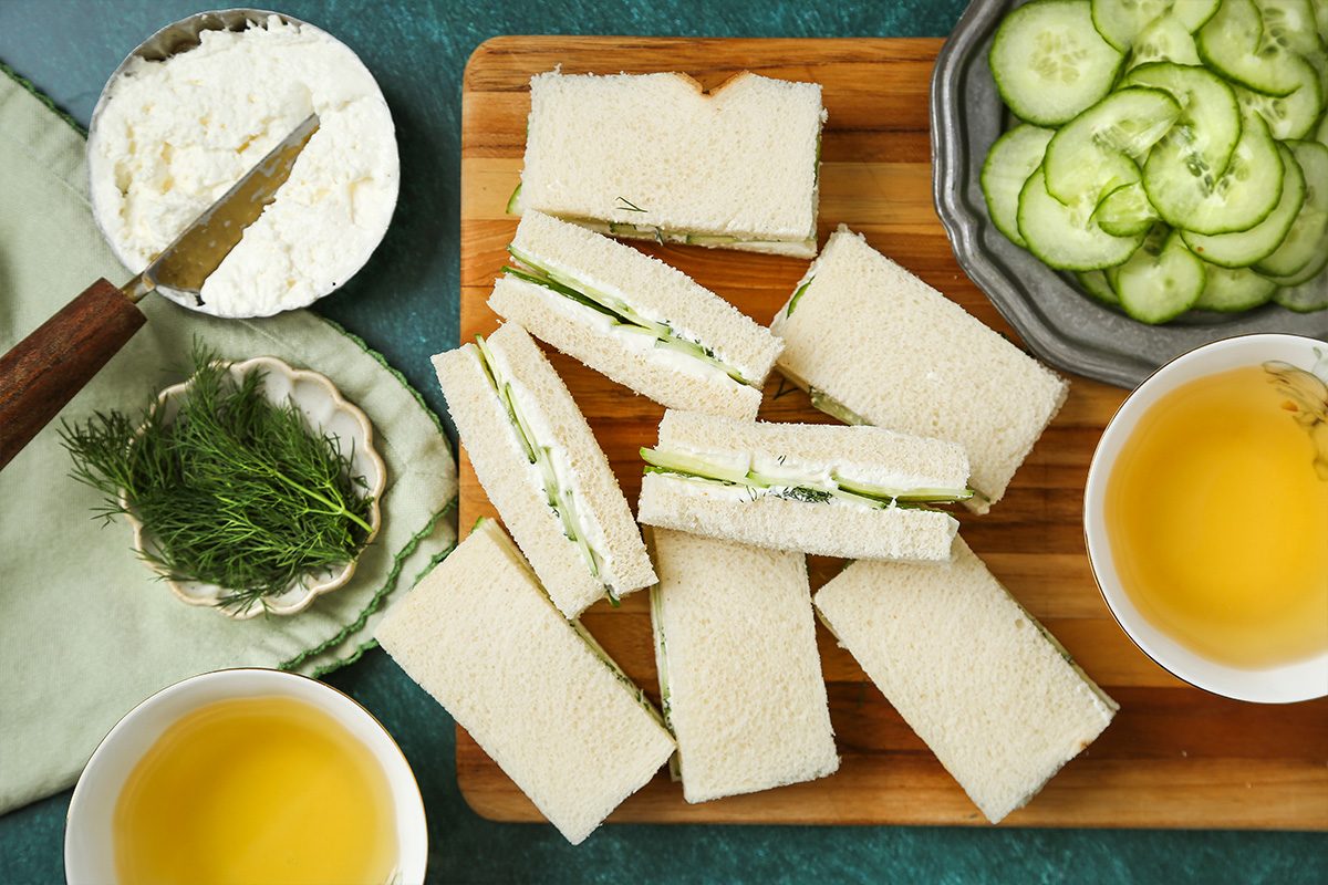 Rectangular cucumber tea sandwiches on a wooden board, surrounded by bowls of cucumber slices, herbs, a dish of soft cheese, and two cups of tea on a green table.