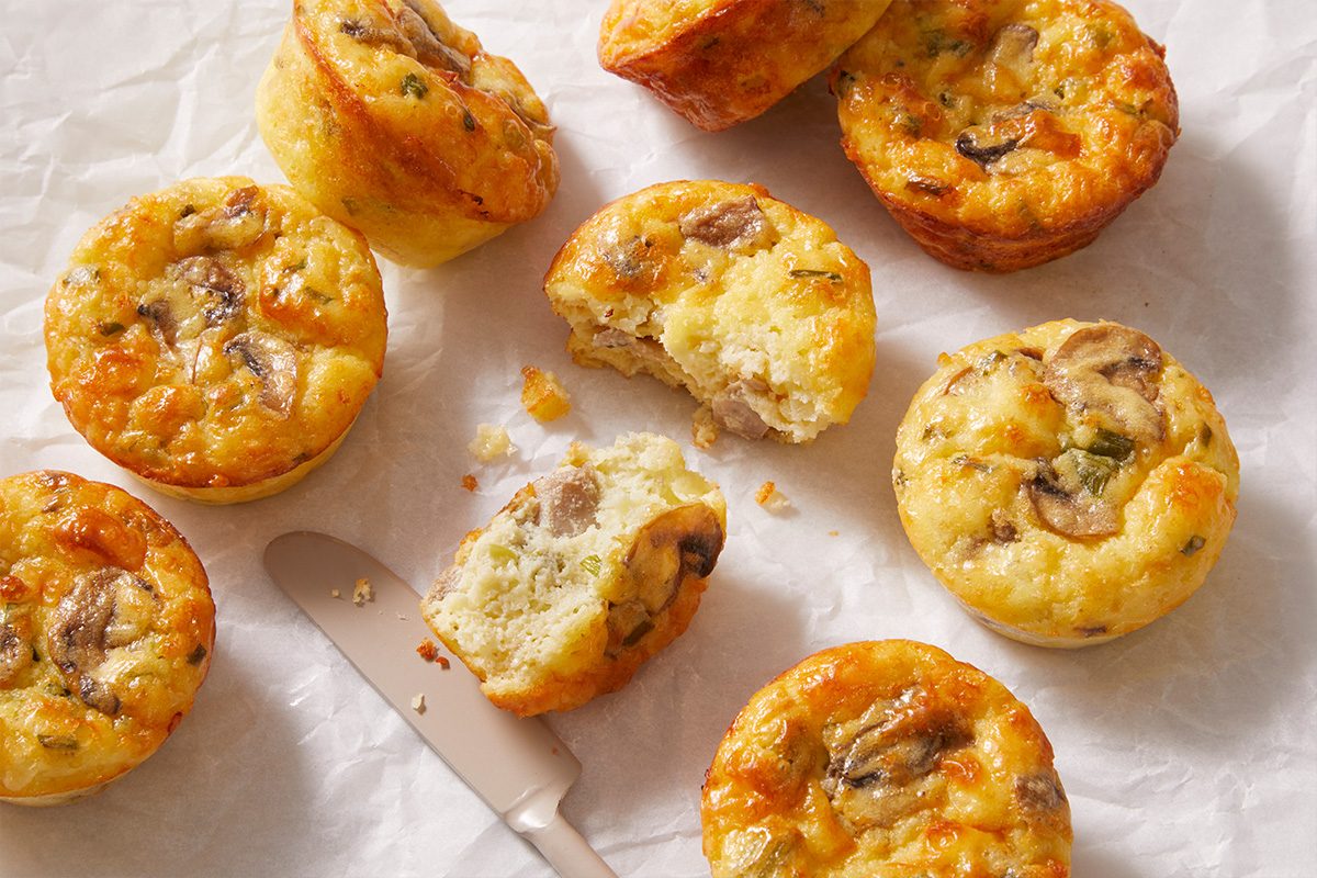 Mini egg muffins with visible pieces of mushroom and herbs are arranged on parchment paper, some whole and one split open with a butter knife nearby, showing their moist, fluffy interior.