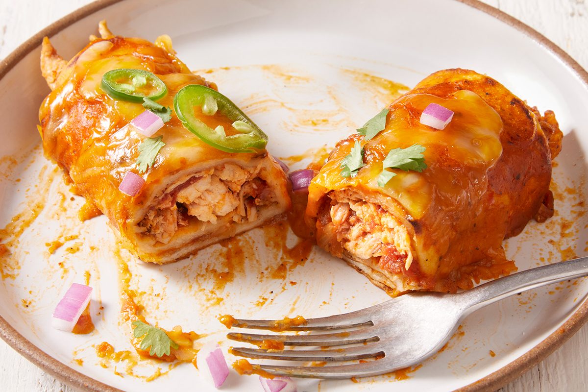 Two enchiladas topped with melted cheese, sliced jalapeños, chopped red onion, and cilantro are cut open on a white plate with a fork, revealing a filling of shredded chicken.