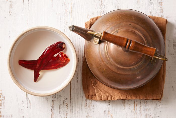 A metal teapot with a wooden handle sits on a folded brown cloth next to a white bowl containing two dried red chili peppers, all placed on a rustic white wooden surface.