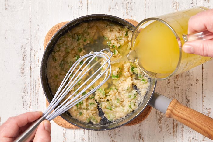 A person whisks a mixture of chopped vegetables and flour in a saucepan while pouring yellow broth into the pan, on a white wooden surface.