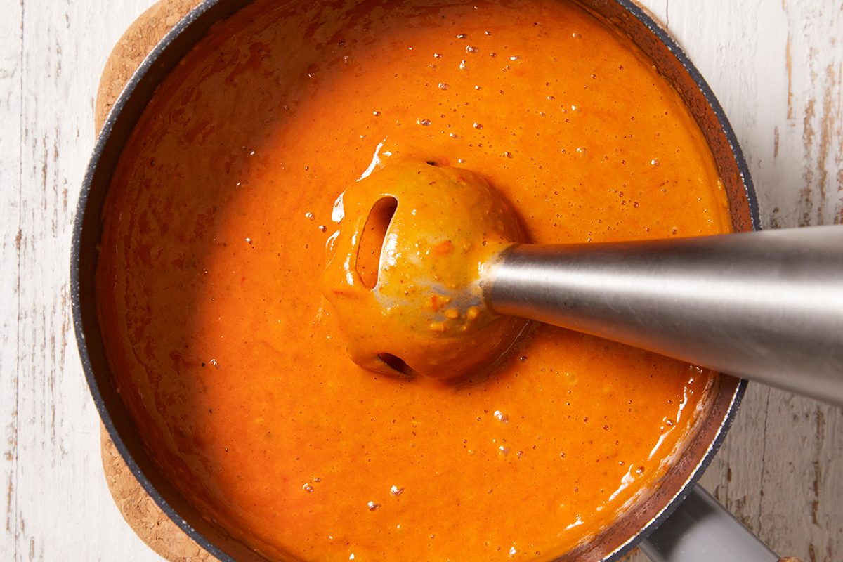 An immersion blender mixes a smooth, bright orange sauce in a pot on a light wooden surface.