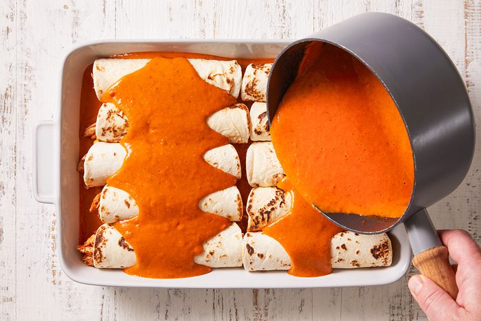 A hand pours bright orange enchilada sauce from a saucepan over rolled tortillas arranged in a white baking dish on a light-colored surface.
