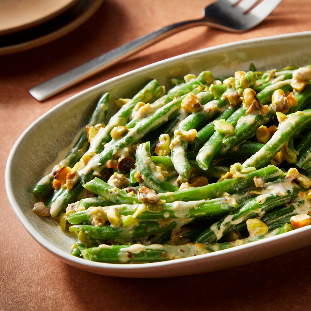 3/4th shot of A serving dish filled with green beans in a creamy sauce, topped with chopped nuts, sits on a brown surface with a fork in the background